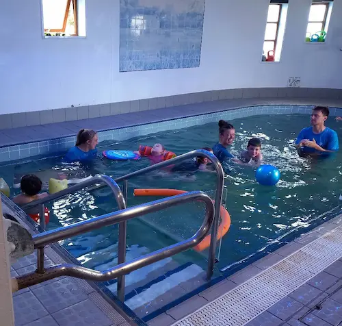 Participants taking part in a supported swimming activity in an indoor pool with instructors and flotation equipment.
