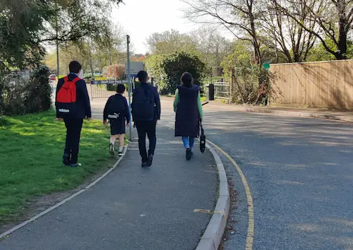 School children and an adult walking along a pavement towards a school entrance.