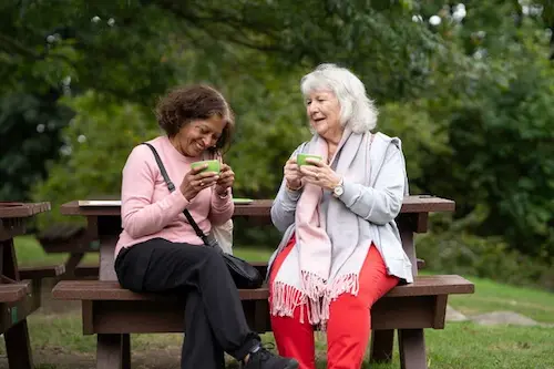 Two ladies sitting at a picnic bench, laughing and holding green tea cups