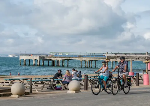 Two people cycling along a seafront promenade beside Boscombe Pier, with benches, beach visitors and the sea in the background.