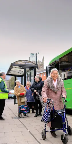 Older people at a bus stop next to a community bus, including a person using a walking aid.