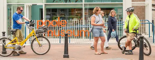 People walking and cycling outside Poole Museum, with bicycles, pavement seating and the museum entrance in the background.
