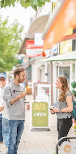 Two people talking on a shopping street, with shopfronts, signs and outdoor seating in the background.
