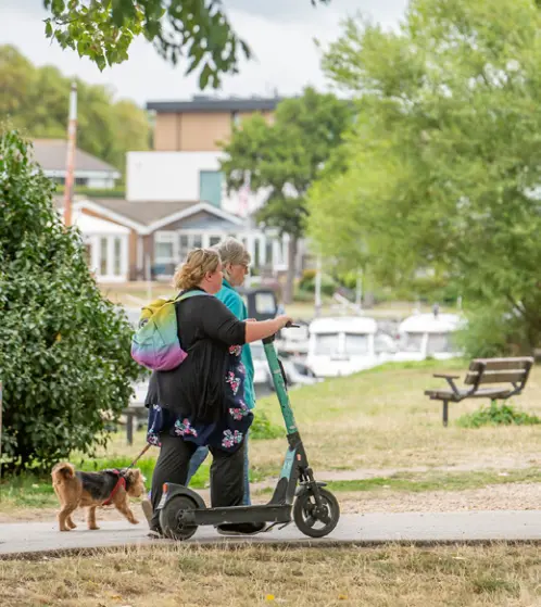 People walking with an e-scooter on a path beside a green space.