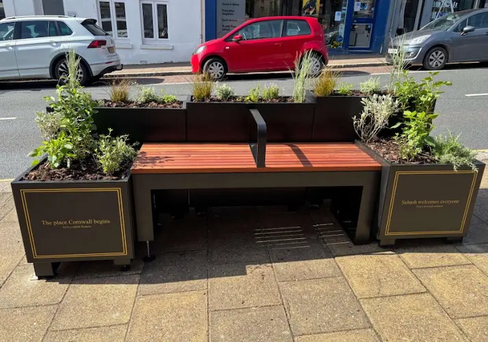 Large rectangular black planter with gold trim containing plants, with a wooden bench built into the centre, located on a street beside parked cars.