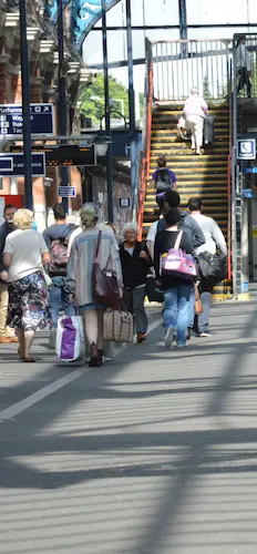 People walking through a busy railway station, with passengers carrying luggage and using stairs to reach the platforms. Station signs and information boards can be seen in the background.