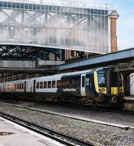 South Western Railway train at a station platform, with railway tracks, station buildings and a glass-roofed structure in the background.
