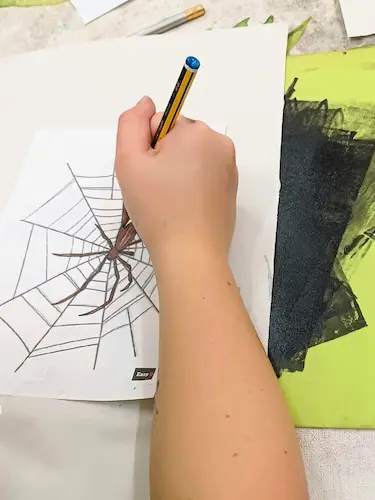 Person drawing a spider on a spider web worksheet with a pencil during an arts and crafts activity.