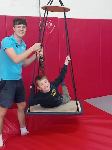 Child using a supported platform swing in an indoor sensory gym with a staff member nearby.