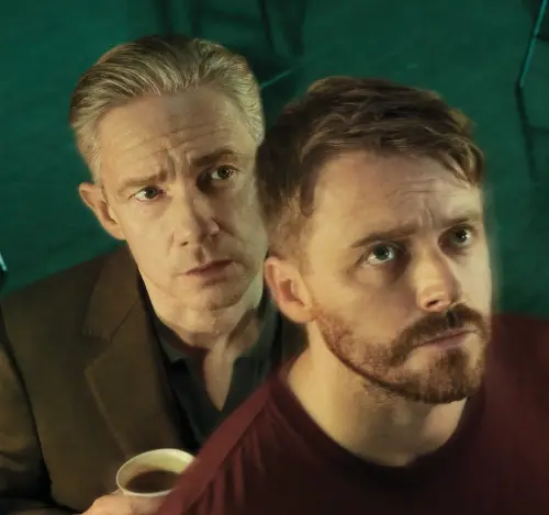 Two people standing close together. The person in the foreground wears a maroon shirt, and the person in the background holds a cup of coffee and wears a dark jacket.