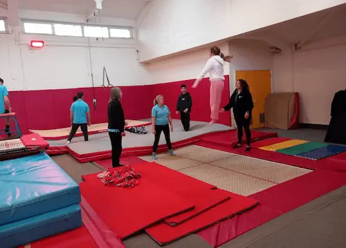Participant bouncing on a trampoline in an indoor gym with staff and other participants around the activity area.