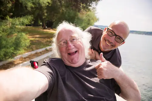 Two people outdoors near a body of water taking a selfie, with one person seated in a wheelchair in the foreground and another person leaning in from behind and giving a thumbs-up, trees and a path visible in the background.