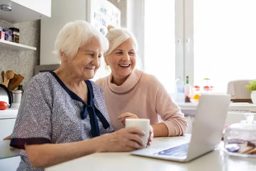 Two people sitting at a kitchen table using a laptop together, with one person holding a mug while both look at the screen, kitchen items and a window in the background.