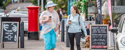 Two people walking and talking along a seaside high street, with a red post box, shopfronts and signs for watersports and boat hire in the background.