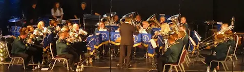 Brass band performing on stage in a semi-circle, with a conductor in the center.