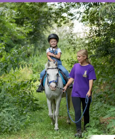 Child riding a pony along a woodland path led by an adult holding the reins.