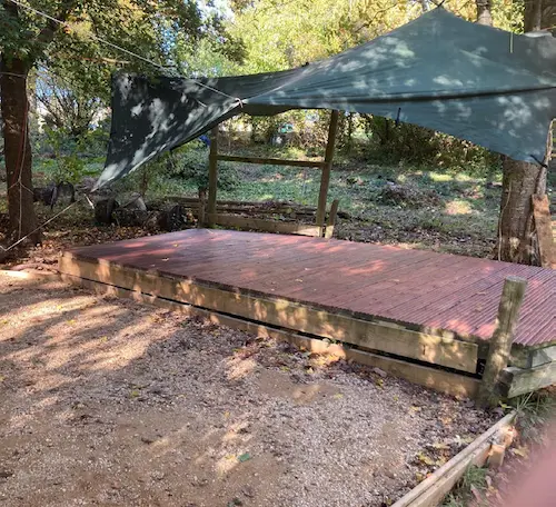 Wooden outdoor stage with overhead shelter canopies, surrounded by trees in a woodland setting.