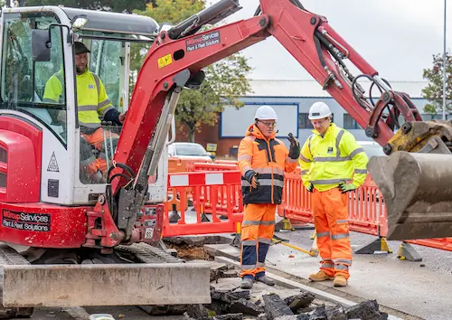 Roadworks taking place beside a road, with construction workers in high-visibility clothing and a digger excavating the pavement behind safety barriers.
