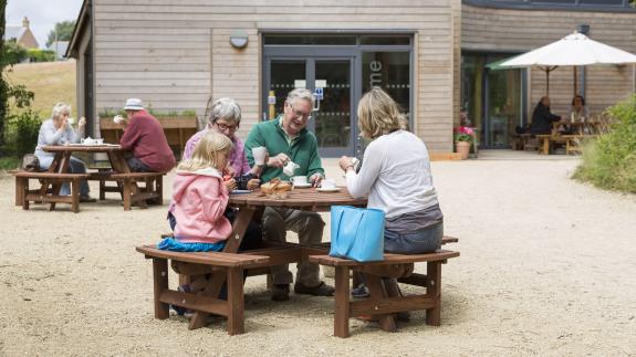family sit on picnic bench in front of visitor centre drinking tea and smiling