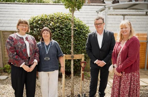 Three white women and one white man stand next to the trunk of a tree which has just been planted in the ground