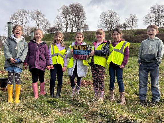children stand in a line in a field smiling with newly planted trees behind them, holding a Nature Recovery Dorset board