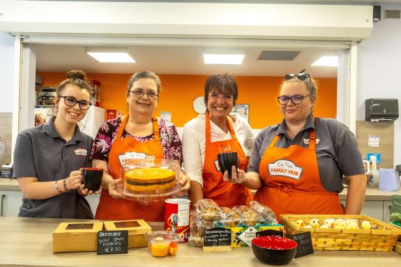 Four women wearing orange aprons are in a cafe holding mugs of coffee and cakes