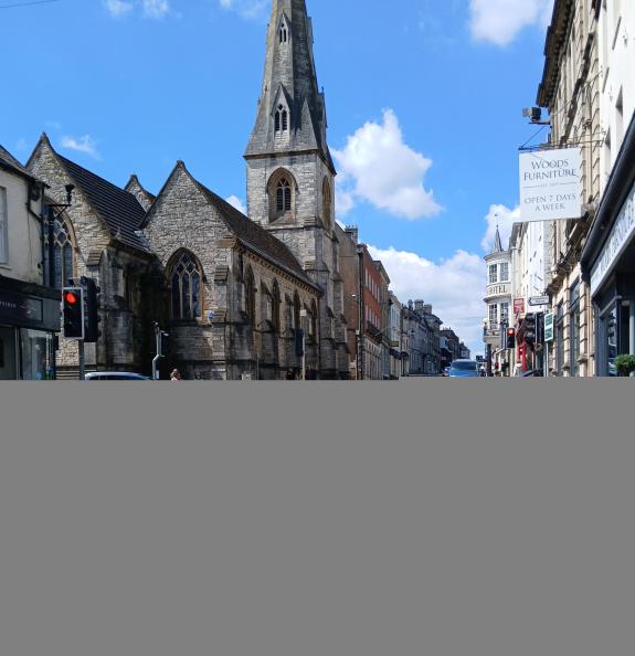 A main road on a sunny day with blue sky, a church in the background and cars waiting at junctions from two side roads. There is a red traffic light and a blue VW camper van waiting at another signal.