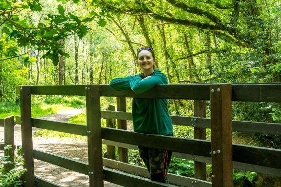 A woman with brown hair and wearing a green fleece and trousers is standing on a bridge surrounded by green trees