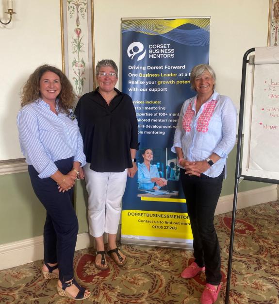 Three smiling people standing by a tall banner that promotes the Dorset Business Mentors service. The two people either side are wearing light blue shirts and the person in the middle is wearing a black top.
