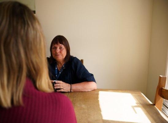 Two women sit at a wooden dining table, facing each other. One woman has dark hair, is wearing a lanyard and holding a mug. The camera is behind another woman's shoulder.