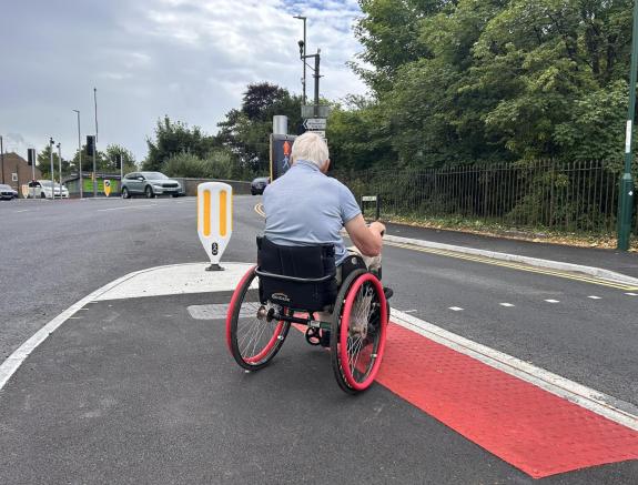 A man seated in a wheelchair on a traffic island waiting to cross a road