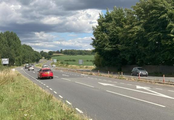 A road with broken white lines. There are arrows on two of the sections and cars, including a red car, a white taxi and a silver car heading in east. There is a grey car travelling west on the other side of the carriageway.