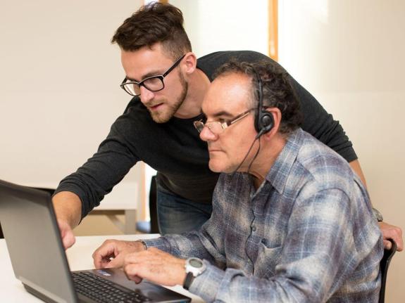 Man at desk with laptop, being helped by another person