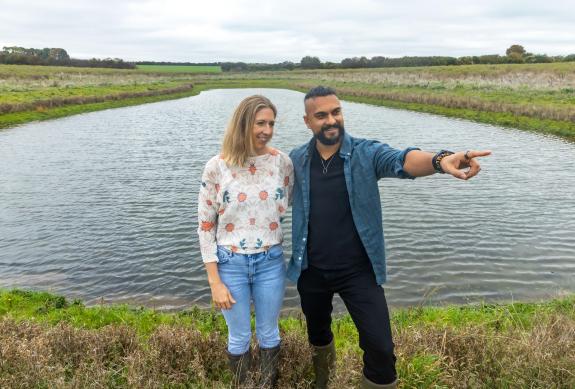 A woman with blonde hair and wearing jeans, wellies and a jumper is standing next to a man with black hair and a black beard, he is wearing a blue shirt, black trousers and wellies. They are both standing in front of a large pond surrounded by countryside