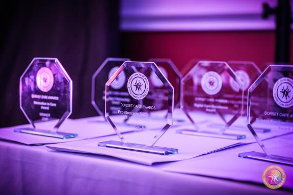 Glass Dorset Care Awards trophies on a table under purple lighting, with the front award reading “Dorset Care Awards” and “Excellence in Social Care Training Award”.