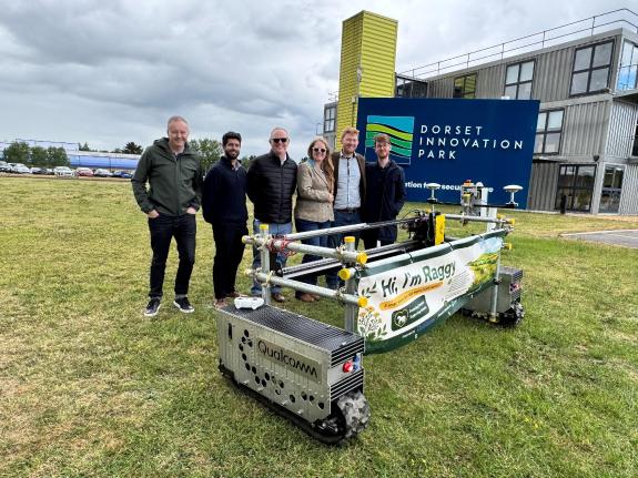 A group of people standing being Raggy the ragwort killer robot and in front of the Dorset Innovation Park sign
