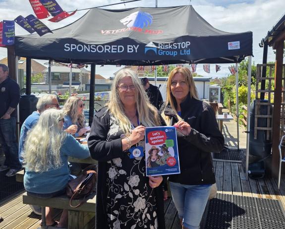 Two people smiling holding a Let's question ageism poster in front of a group of people laughing while seated at a table
