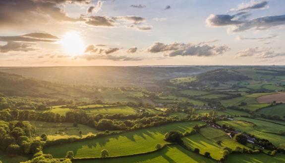 Sunset over rolling green countryside with patchwork fields, hedgerows and a small village in a valley, long shadows stretching across the landscape beneath a partly cloudy sky.