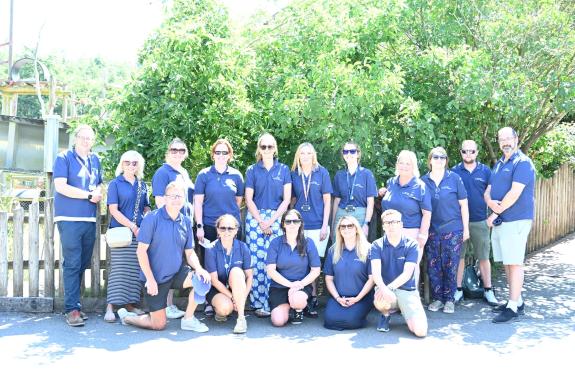 A group of around 16 adults wearing matching navy-blue polo shirts pose for a group photo outdoors on a sunny day. Some people are standing while others are kneeling in front. The group is gathered on a paved path in front of a wooden fence.