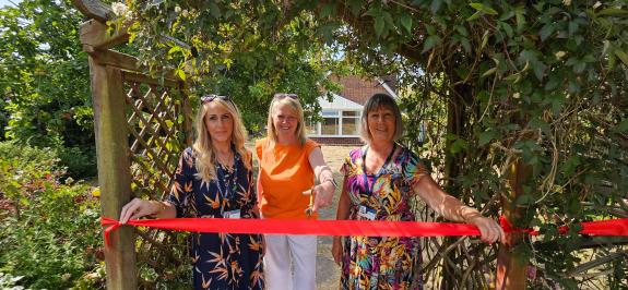 Three women stand behind a taut red ribbon which is attached to a wooden archway in a back garden. The middle woman has a pair of ceremonial scissors.