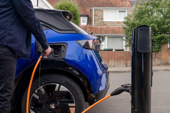 A blue electric vehicle is being charged up by a man using an orange hose, on the roadside.