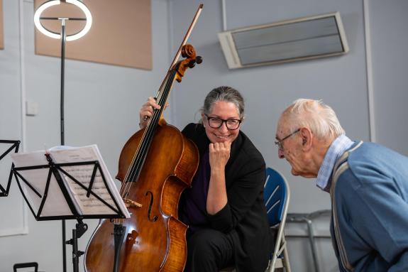 A female middle-aged double base player is sat down with her instrument in front of her. She is looking to her left at an elderly man who is stood up.