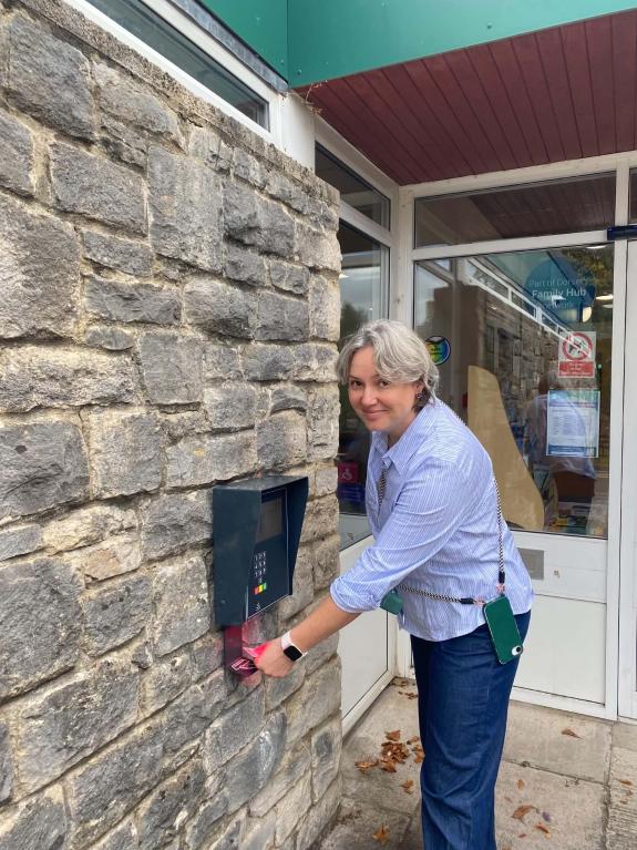 White woman with grey hair and wearing blue clothes is looking at the camera whilst scanning a library card under a reader which is on an external old stone wall, next to the library entrance.