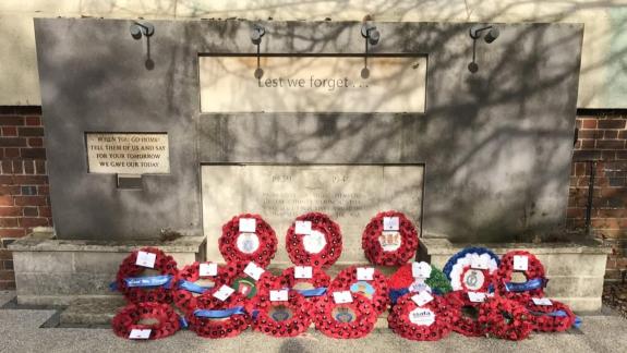 War memorial with the inscription “Lest we forget” and a commemorative plaque, surrounded by numerous red remembrance wreaths laid at its base. The memorial stands against a stone wall with tree shadows cast across the surface, and wreaths.