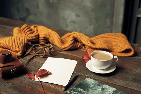 Various autumnal toned objects on a wood table. Includes a mustard yellow scarf, red autumn leaves and a cup of coffee.