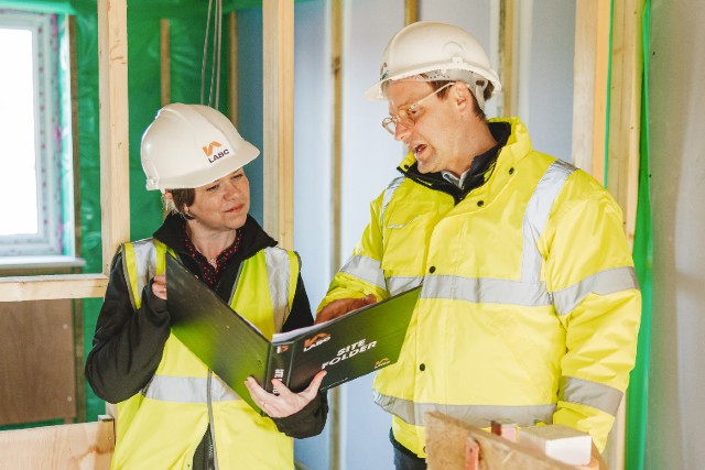 A woman and a man stand in a building site, wearing high-vis and hard hats, looking at some plans.